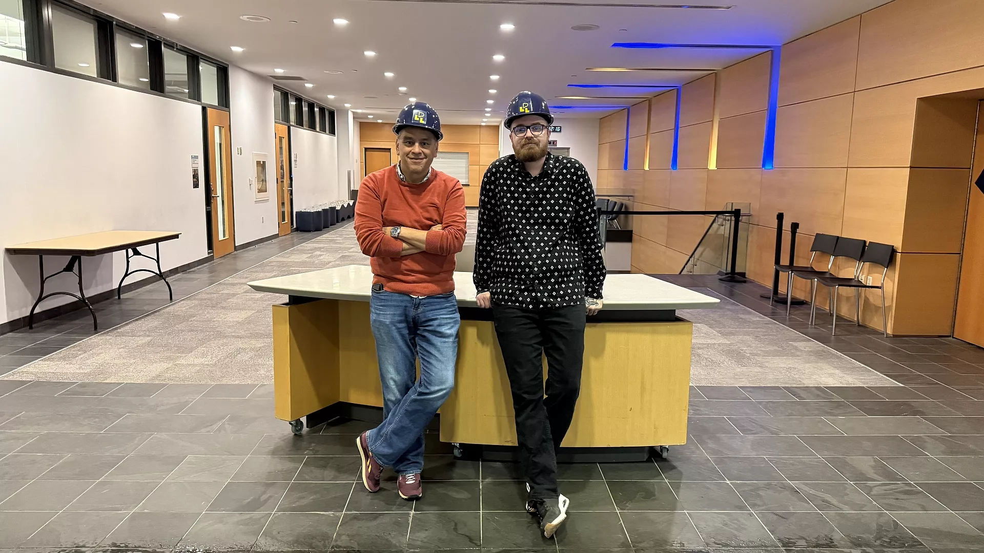 Photo of two members of the Wolf Performance Hall staff team standing in front of the Crush Space area before the renovation. They are wearing hardhats with the Library logo and smiling.