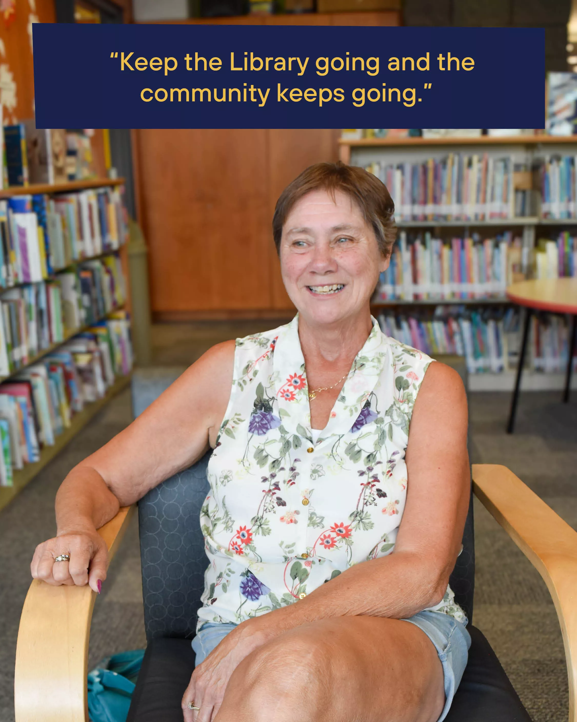 A smiling woman sits in a chair in the library with bookshelves in background. Text reads Keep the Library going and the community keeps going.